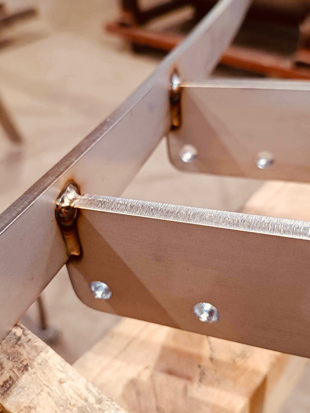 Fragment metalowej konstrukcji z widocznymi spawami i nitami, leżącej na drewnianym podłożu, uchwycony z bliska pod kątem.