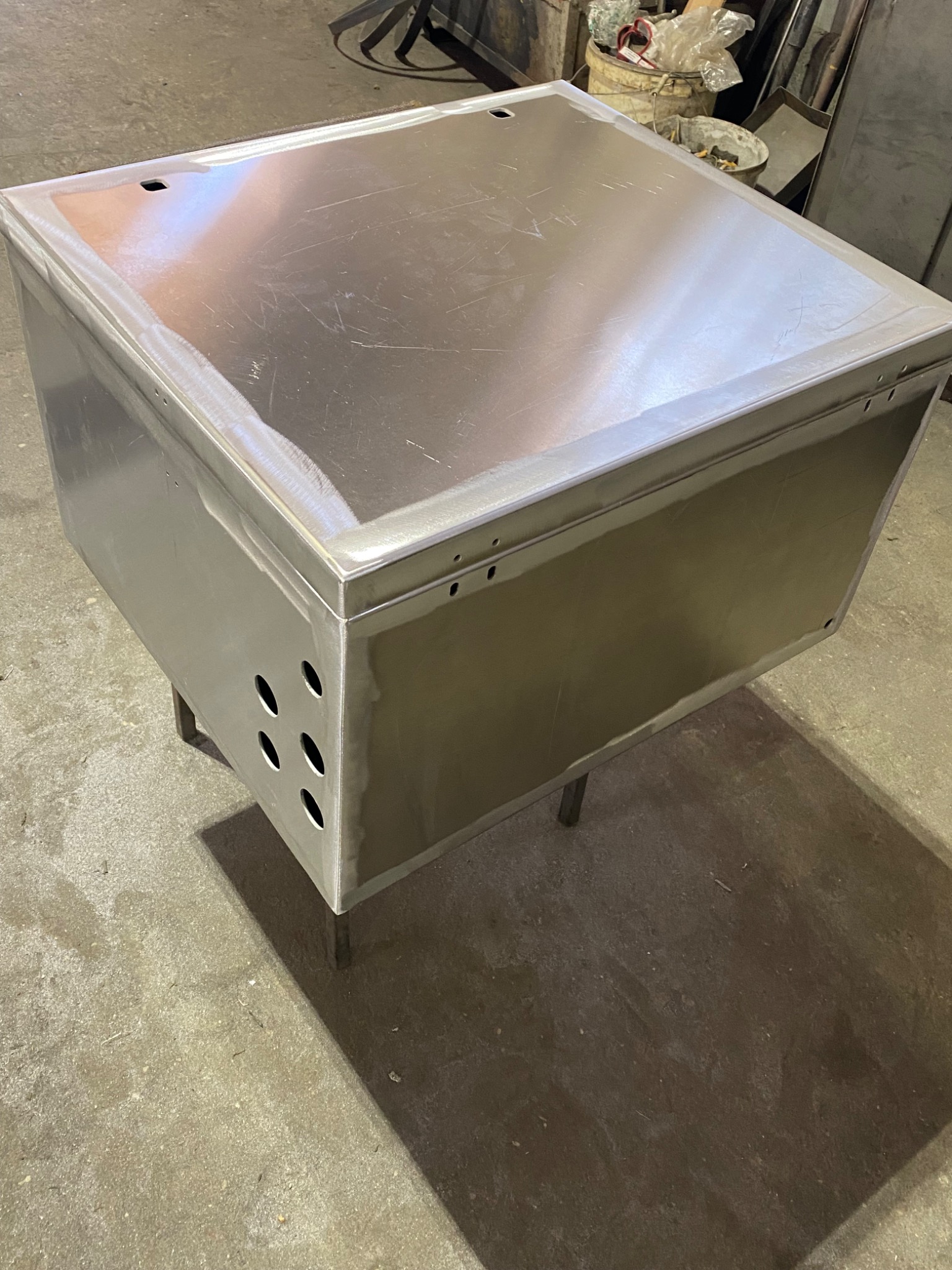 Metalowa skrzynia na czterech nogach, wykonana ze stali, z otworami wentylacyjnymi po bokach, widoczna na betonowej posadzce.