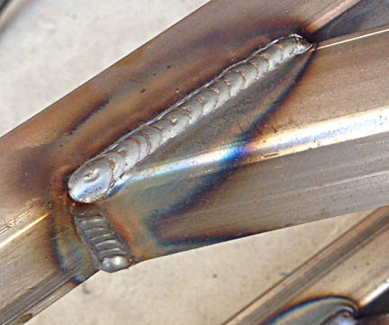 Spoina łącząca dwa metalowe profile pod kątem, widoczne ślady ciepła i tęczowe przebarwienia na metalu.
