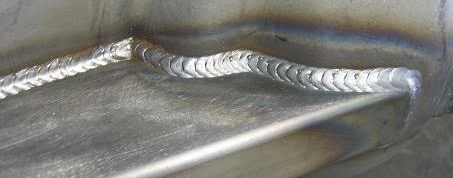 Zbliżenie na spoinę wykonaną metodą MIG/MAG na metalowej powierzchni, widoczne charakterystyczne ślady po spawaniu w kształcie półksiężyców, z lekkimi przebarwieniami termicznymi na materiale.