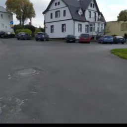 Dwupiętrowy dom z białymi ścianami i ciemną dachówką, widoczny z ulicy z zaparkowanymi samochodami, drzewem i fragmentem sąsiedniego budynku.