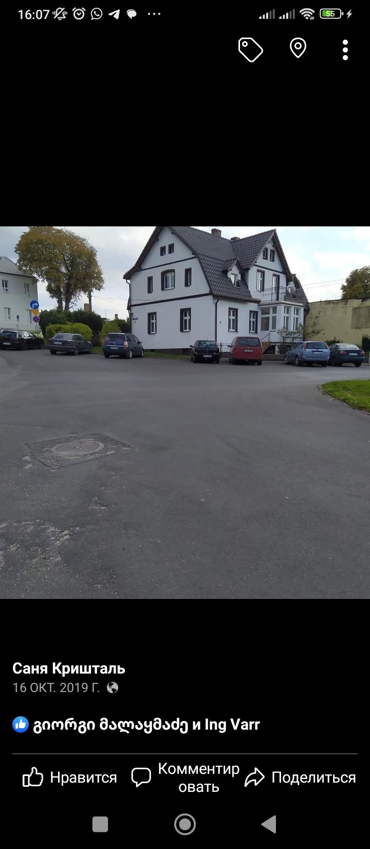 Dwupiętrowy dom z białymi ścianami i ciemną dachówką, widoczny z ulicy z zaparkowanymi samochodami, drzewem i fragmentem sąsiedniego budynku.