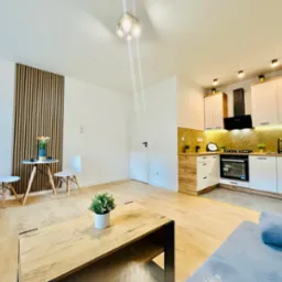 Jasne, przestronne wnętrze mieszkania po remoncie: drewniana podłoga, białe ściany, nowoczesna zabudowa kuchenna z drewnianymi akcentami i czarnym okapem, stół z krzesłami oraz dekoracyjny panel...