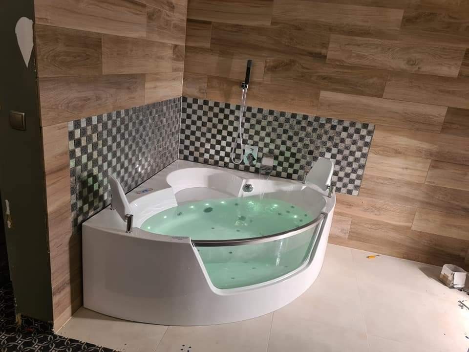 Narożna wanna z hydromasażem i szklanym panelem, wypełniona wodą z zielonym podświetleniem, otoczona płytkami w szachownicę i drewnopodobnymi panelami w łazience w Rabce-Zdrój.