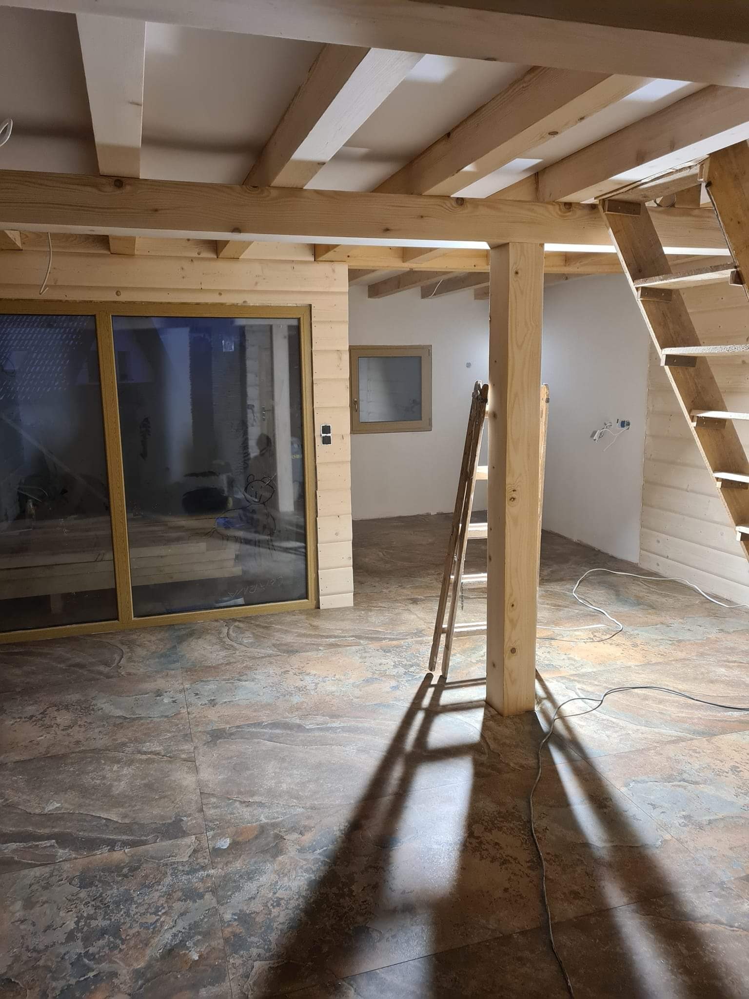 Wnętrze domu w trakcie prac wykończeniowych: widoczne drewniane belki stropowe, schody, okno przesuwne z rysunkami dziecięcymi, drabina oparta o drewniany słup i płytki podłogowe imitujące kamień.