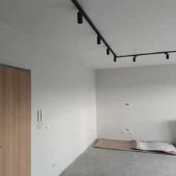 MAZUR BUD Oleksandr Mazur - Wnętrze w trakcie prac przygotowawczych: surowa podłoga, zabezpieczona tektura, białe ściany z widocznymi gniazdkami i włącznikami, nowoczesne czarne oświetlenie szynowe sufitowe.