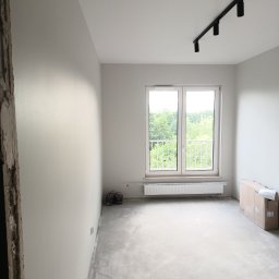 MAZUR BUD Oleksandr Mazur - Wnętrze pokoju po malowaniu ścian na biało, widoczne okno z widokiem na zieleń, grzejnik, lampa sufitowa na szynie, podłoga w trakcie przygotowania, kartony w rogu i puszki z farbą przy ścianie.