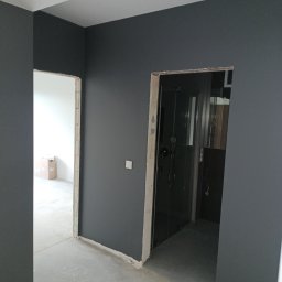 MAZUR BUD Oleksandr Mazur - Pomieszczenie w trakcie prac wykończeniowych, ściany pomalowane na ciemny grafit, widoczne surowe ościeżnice drzwiowe, w tle kabina prysznicowa i kartony.