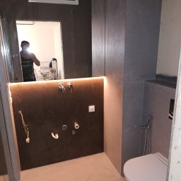 MAZUR BUD Oleksandr Mazur - Wnętrze łazienki w trakcie prac wykończeniowych, widoczne szare płytki na ścianach, podświetlona wnęka z wyprowadzonymi instalacjami wodnymi, toaleta wisząca oraz odbicie osoby w lustrze.