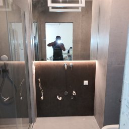 MAZUR BUD Oleksandr Mazur - Szara łazienka w trakcie prac wykończeniowych, widoczne przygotowane przyłącza wodno-kanalizacyjne, częściowo zamontowana kabina prysznicowa i toaleta, oraz odbicie osoby robiącej zdjęcie w lustrze.