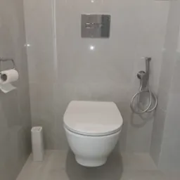 Nowoczesna toaleta wisząca z bidetką w łazience wykończonej szarymi płytkami imitującymi marmur, z widocznym pojemnikiem na zapas papieru toaletowego i puszką farby Dulux na górnej półce.