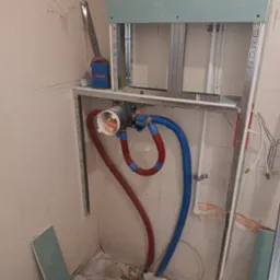 Przygotowanie instalacji wodnej i stelaża pod zabudowę z płyty karton-gips w narożniku łazienki, widoczne rury w izolacji czerwonej i niebieskiej, profil metalowy, fragment płyty gipsowej opartej...