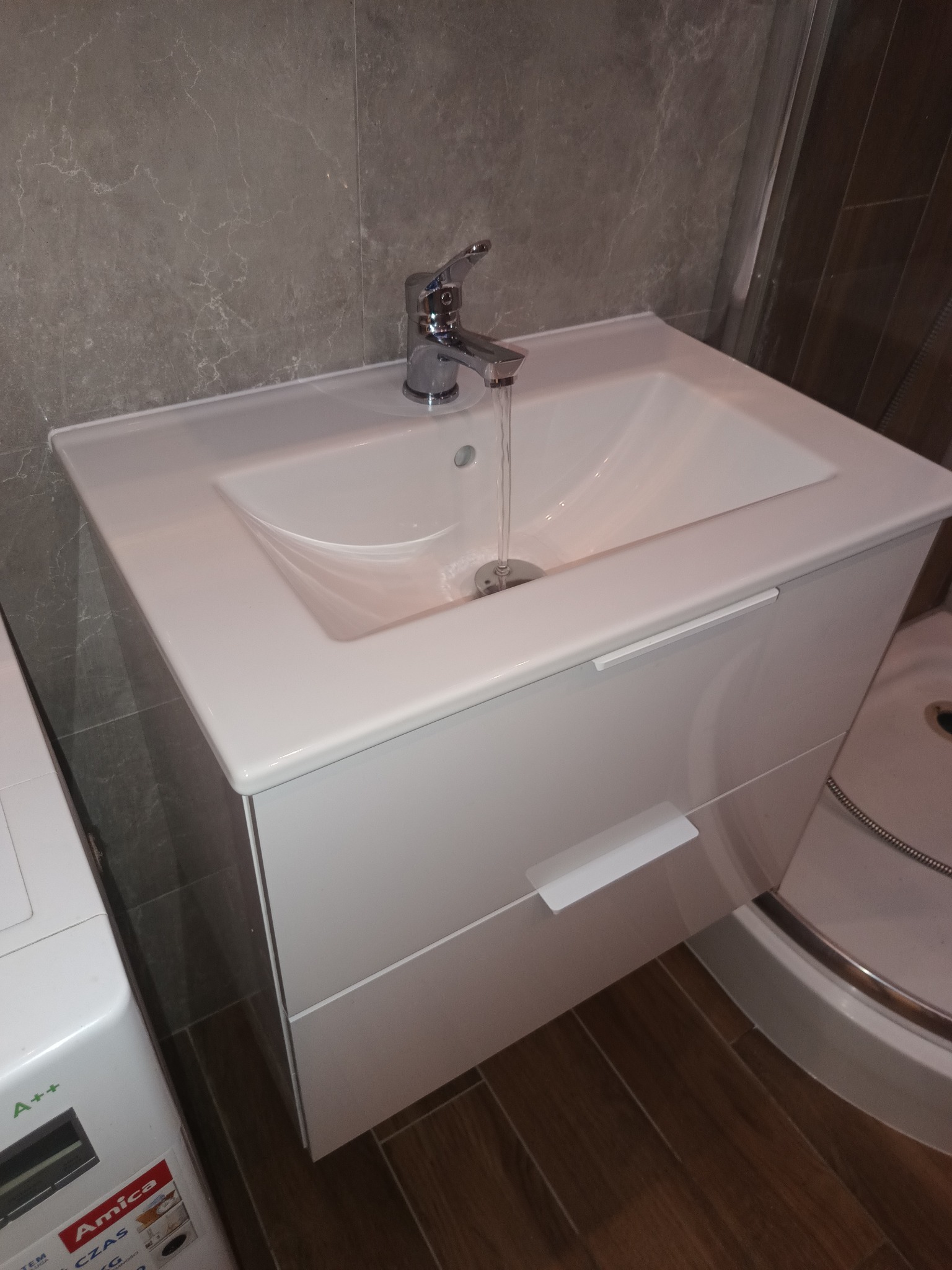 Biała umywalka z szafką i chromowaną baterią, z której leci woda, na tle szarej ściany z płytek imitujących marmur, obok widoczny fragment pralki i kabiny prysznicowej.