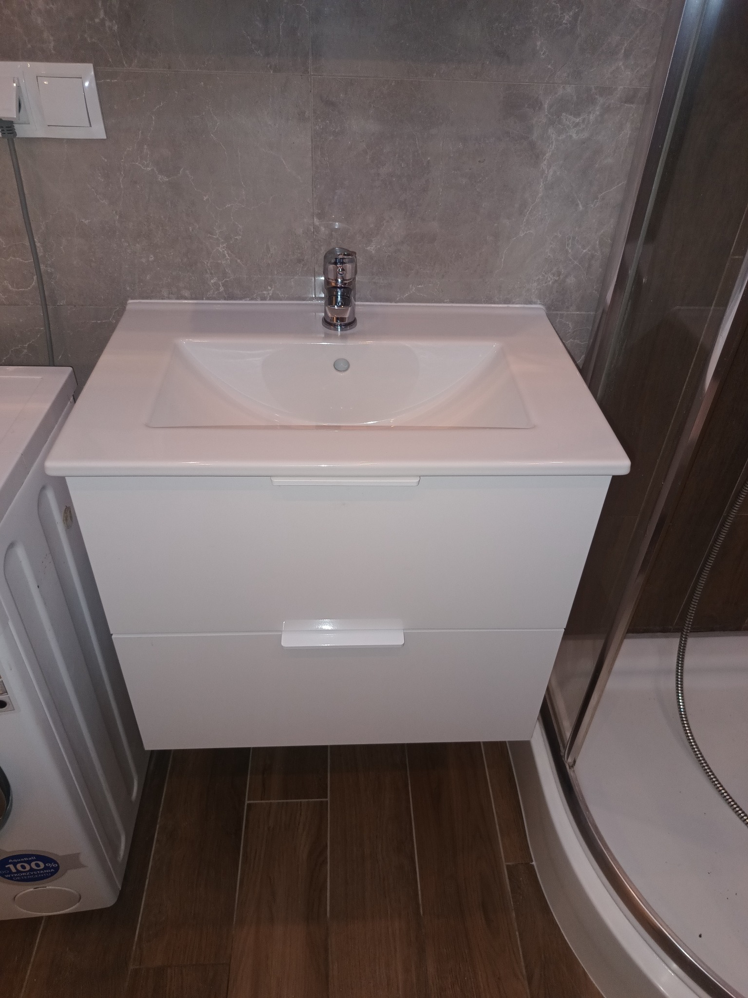 Nowoczesna, biała szafka pod umywalkę z dwiema szufladami i chromowaną baterią, na tle szarych płytek imitujących marmur, obok kabiny prysznicowej i pralki.