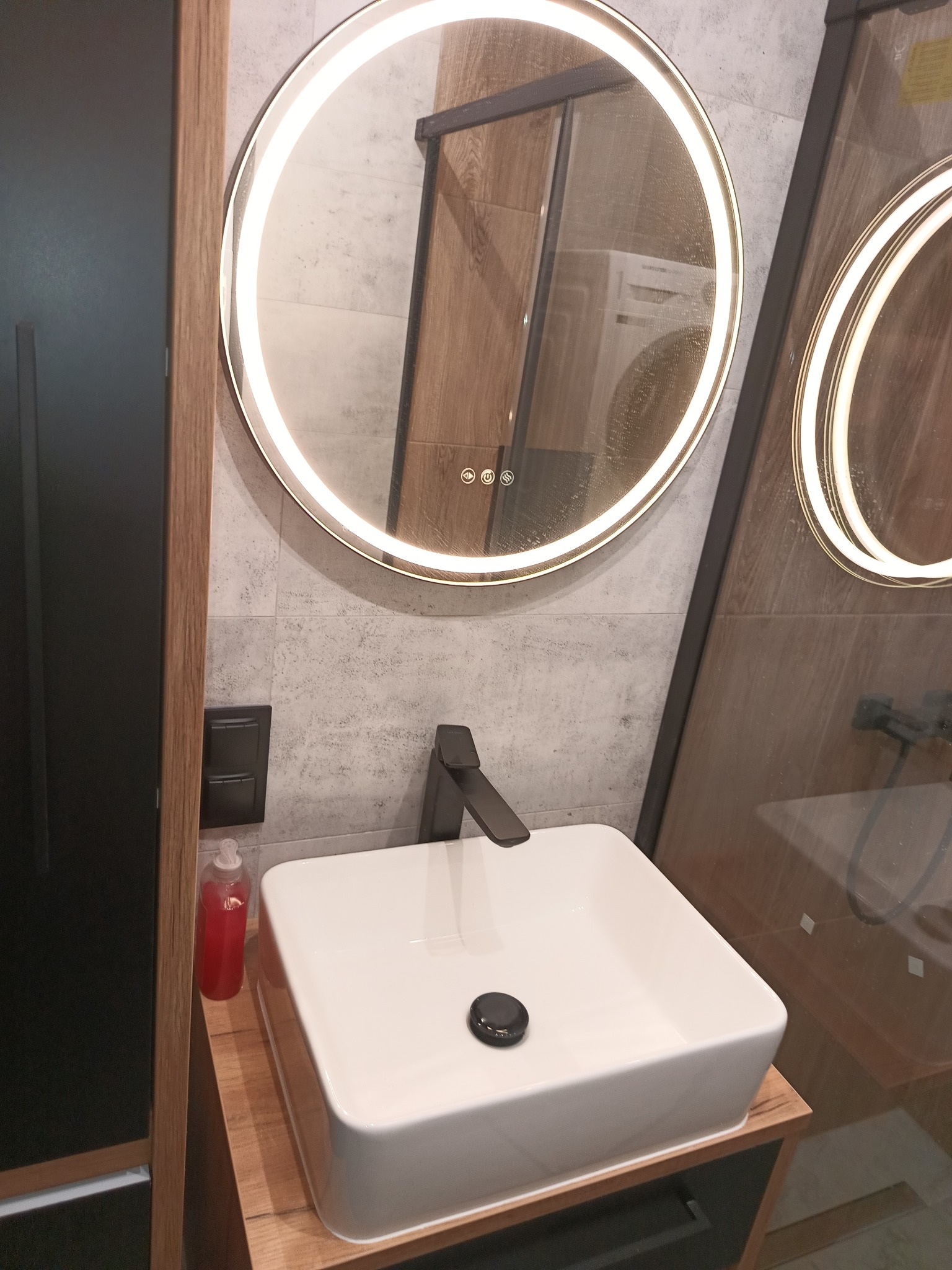 Nowoczesna łazienka z kwadratową umywalką nablatową, czarną armaturą i okrągłym lustrem z podświetleniem LED, odbijającym pralkę w tle.