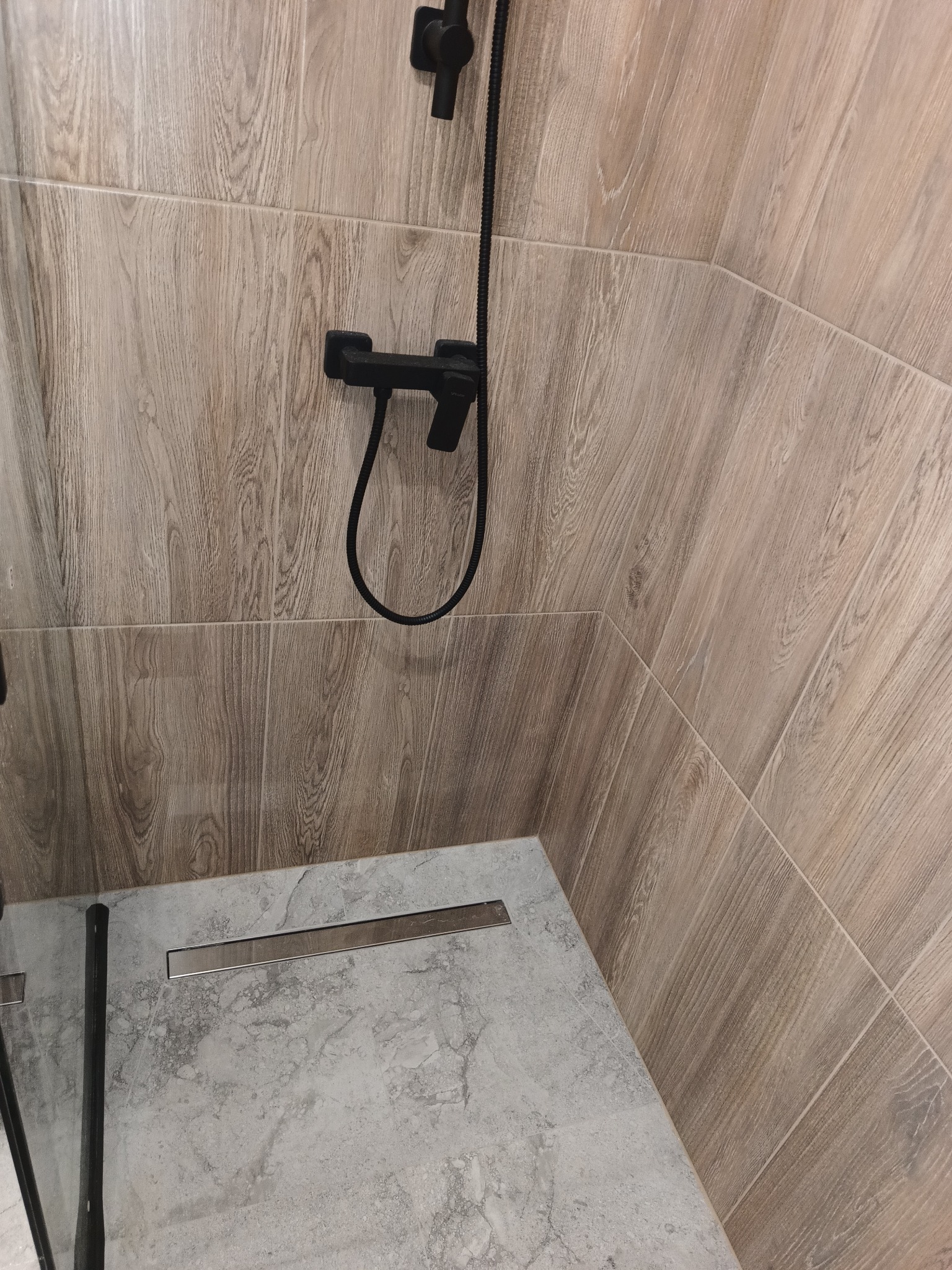 Wykończona kabina prysznicowa z odpływem liniowym i baterią w kolorze czarnym, ściany wyłożone płytkami imitującymi drewno.