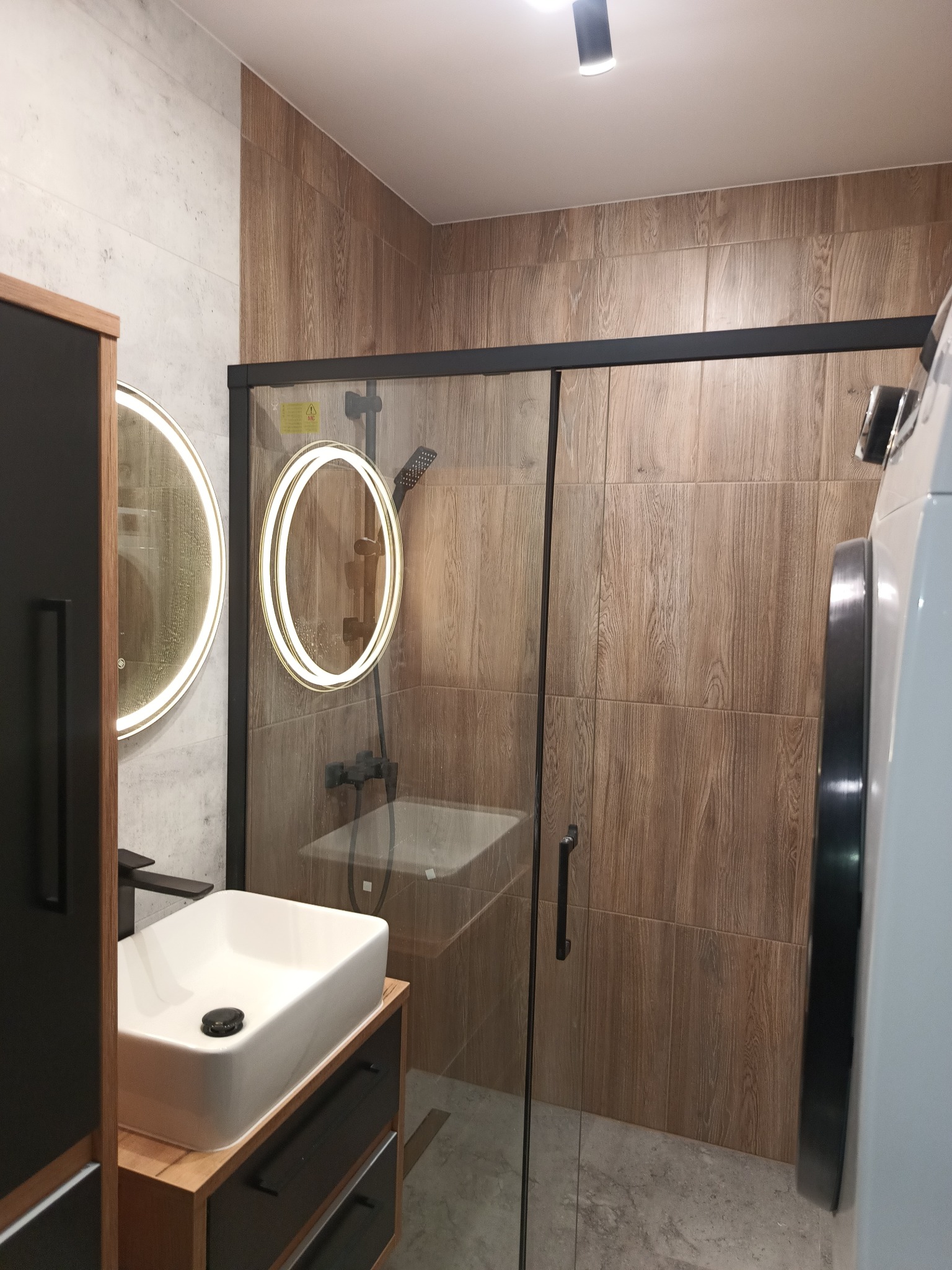 Nowoczesna łazienka z drewnopodobnymi płytkami, kabiną prysznicową ze szklanymi drzwiami przesuwnymi, umywalką nablatową na szafce z czarnymi frontami i okrągłymi lustrami podświetlanymi LED.