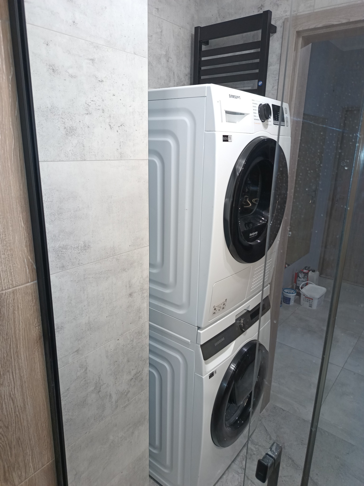 Ustawiona w pionie pralka i suszarka Samsung w łazience wykończonej szarą, imitującą kamień płytką, widoczny grzejnik drabinkowy i fragment szklanej kabiny prysznicowej.