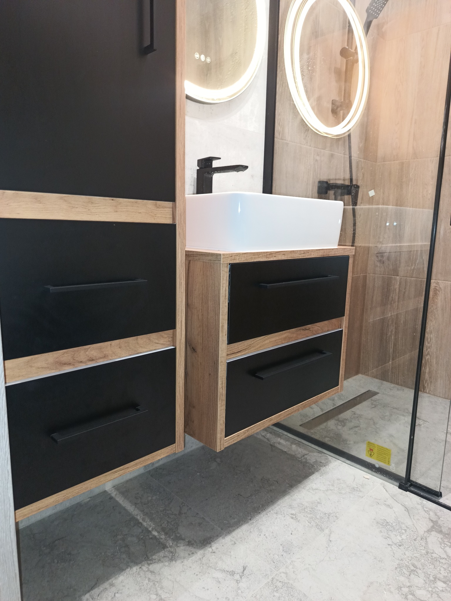 Nowoczesna łazienka z czarnymi szafkami z drewnianymi akcentami, białą umywalką nablatową i okrągłymi lustrami z podświetleniem LED, widoczna kabina prysznicowa z odpływem liniowym.