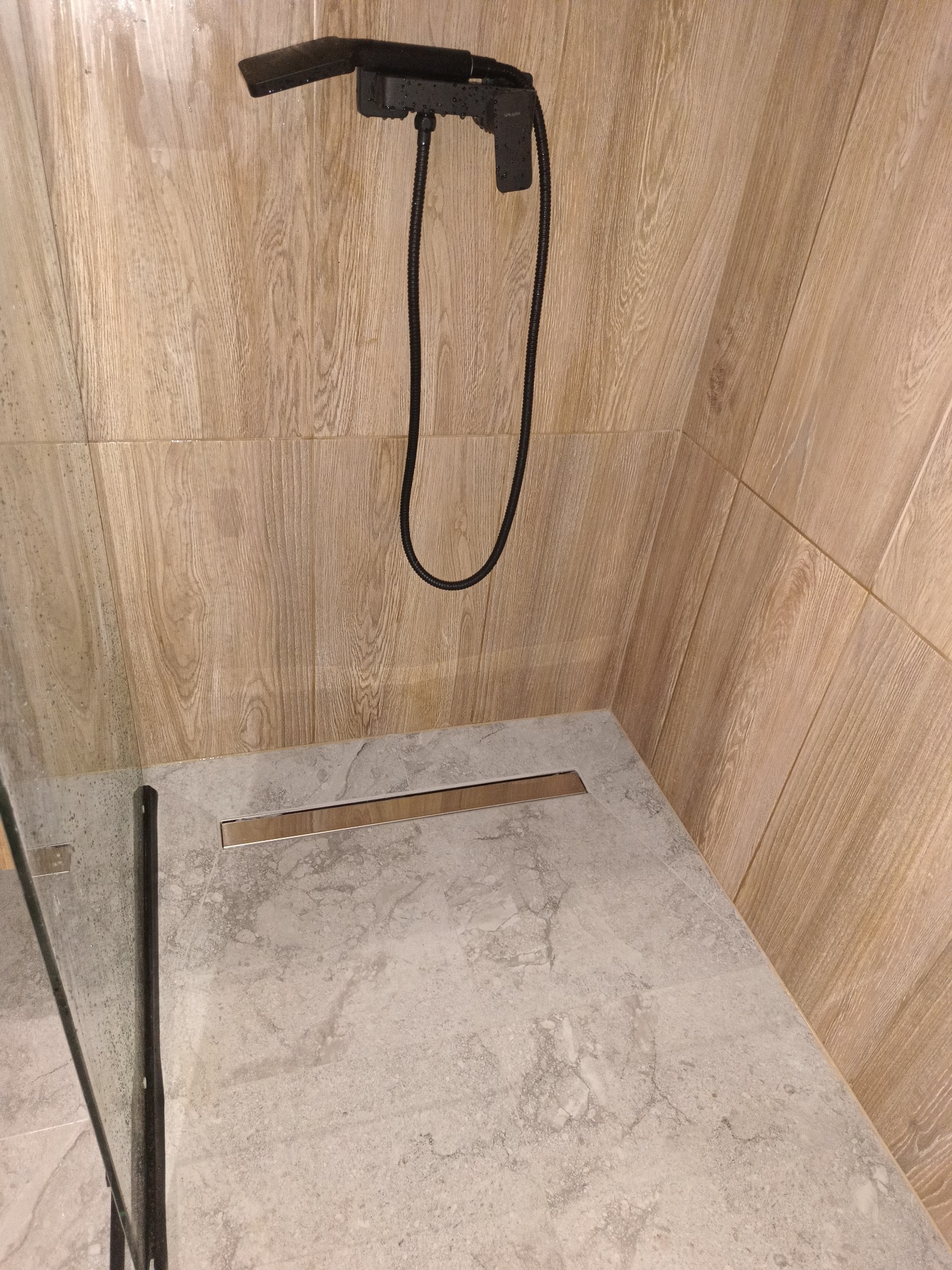 Nowoczesna kabina prysznicowa z deszczownicą w kolorze czarnym, wykończona płytkami imitującymi drewno na ścianach i szarym marmurem na podłodze z odpływem liniowym.