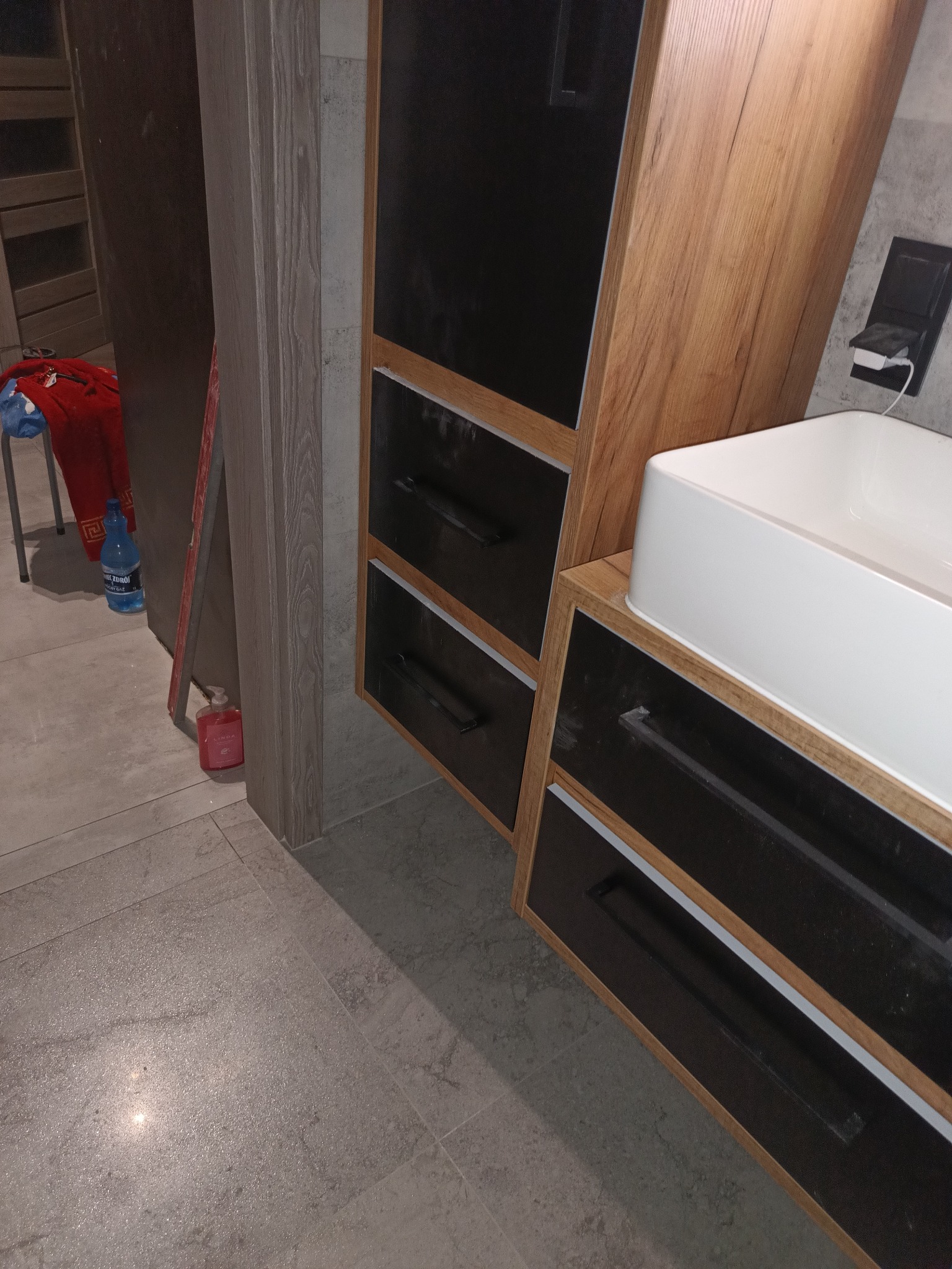 Nowoczesna szafka łazienkowa z umywalką nablatową, czarne fronty szuflad z długimi uchwytami, drewnopodobny korpus, widoczne ślady kurzu na powierzchniach, butelka wody mineralnej w tle.