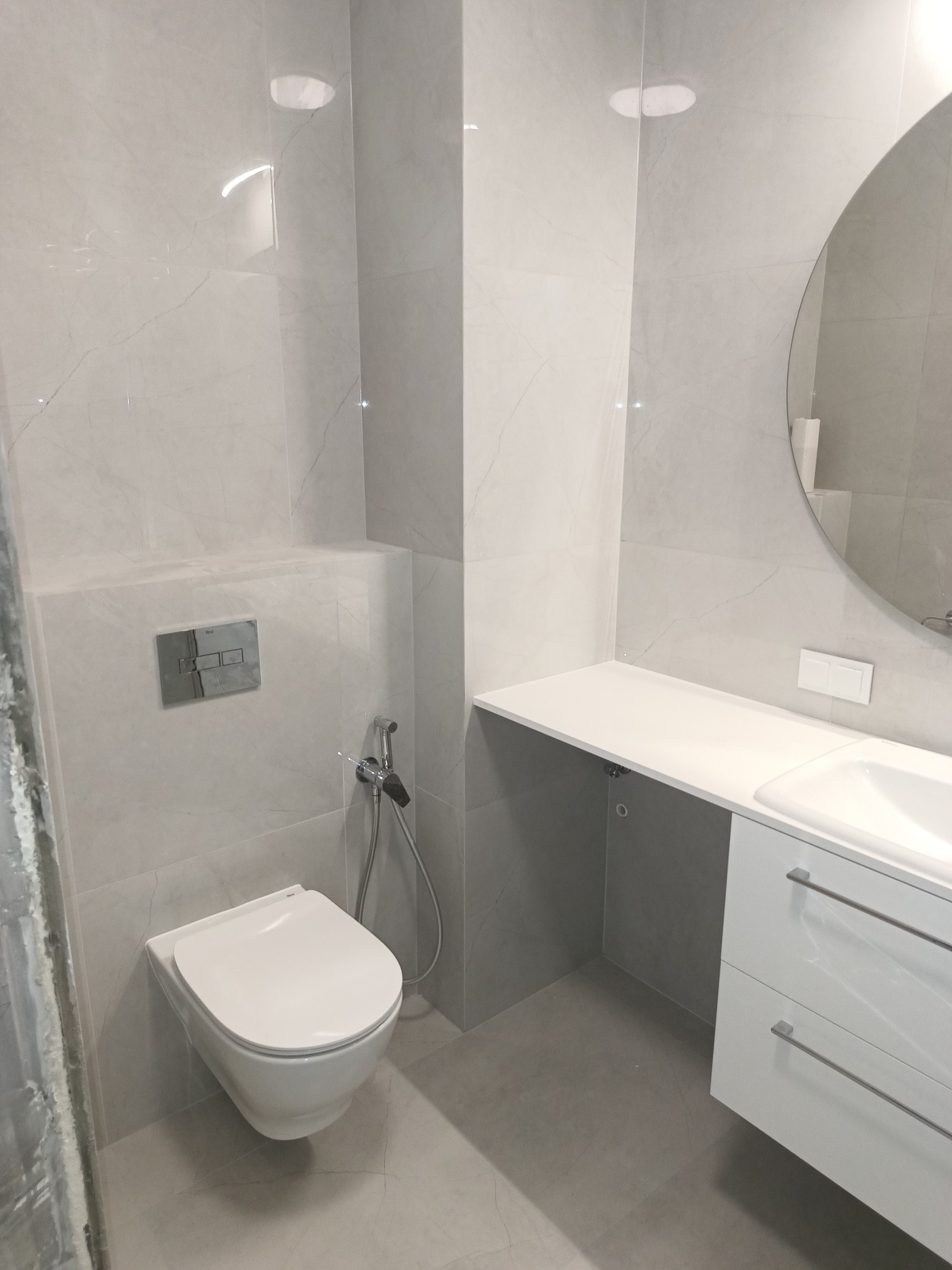 Nowoczesna, minimalistyczna łazienka w odcieniach szarości z wiszącą toaletą i umywalką z szafką, okrągłe lustro na ścianie wyłożonej płytkami imitującymi marmur.