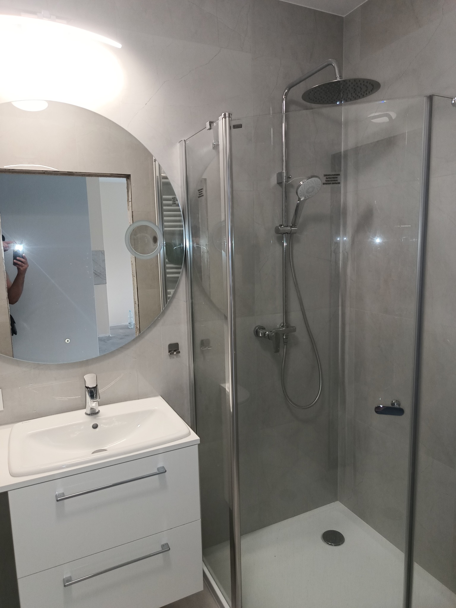 Nowoczesna łazienka w odcieniach szarości z kabiną prysznicową typu walk-in, okrągłym lustrem z podświetleniem i białą szafką pod umywalką z chromowanymi uchwytami, widoczne odbicie osoby robiącej...