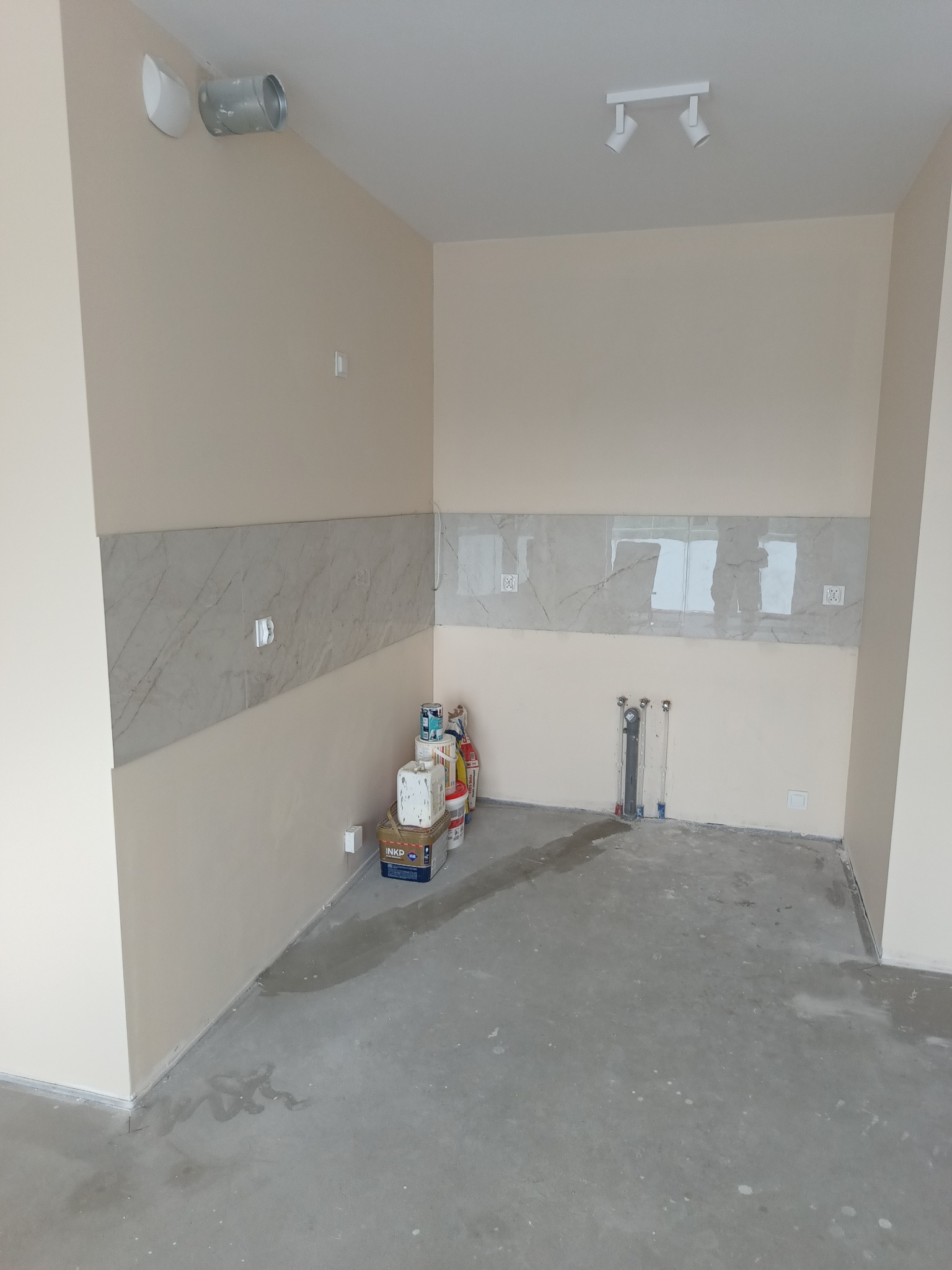 Wnętrze w trakcie prac wykończeniowych: częściowo położone marmurowe płytki na ścianie, widoczne puszki z farbami i gładzią, surowy beton na podłodze, instalacja wodno-kanalizacyjna bez armatury.