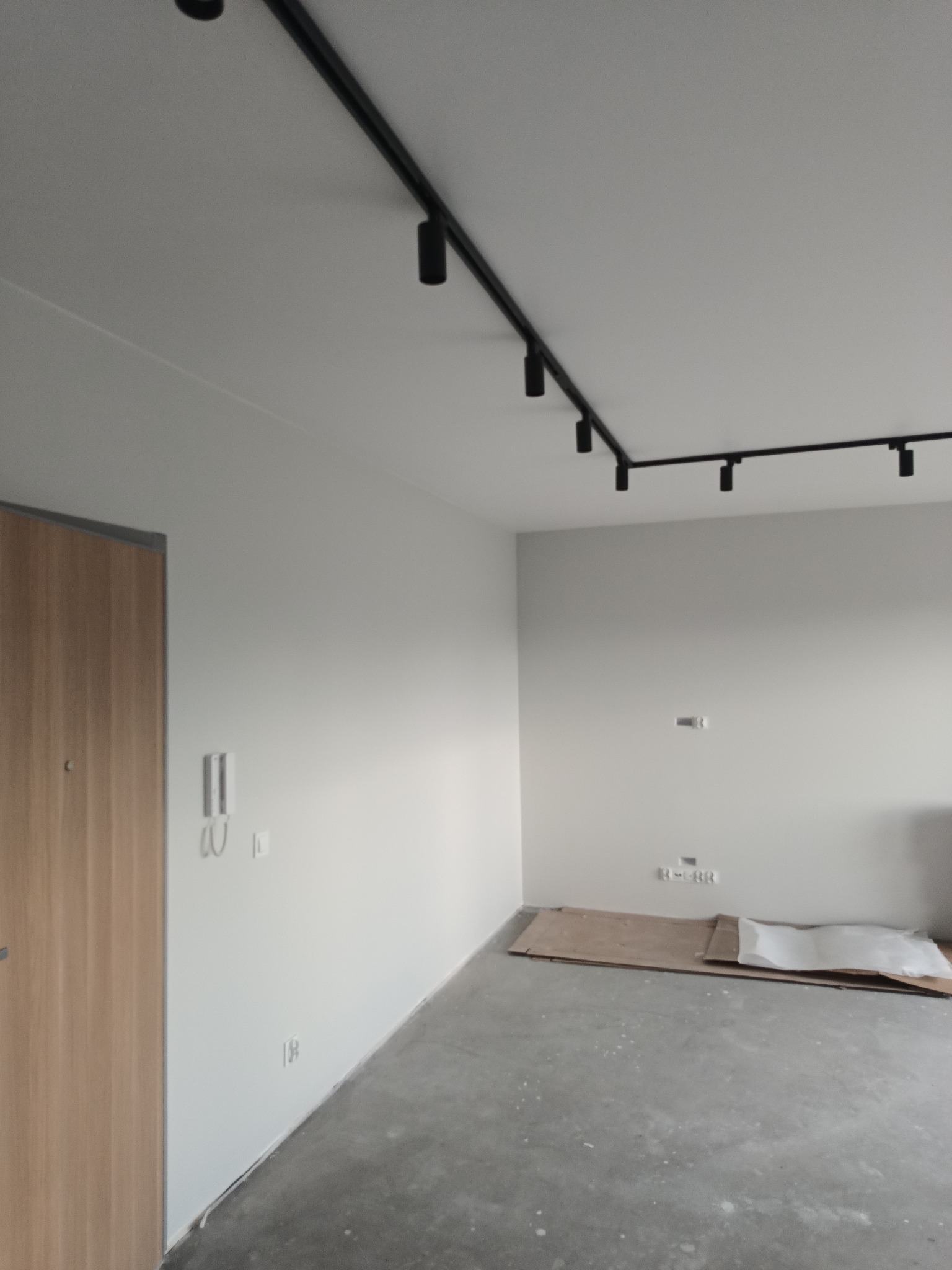 Wnętrze w trakcie prac przygotowawczych: surowa podłoga, zabezpieczona tektura, białe ściany z widocznymi gniazdkami i włącznikami, nowoczesne czarne oświetlenie szynowe sufitowe.