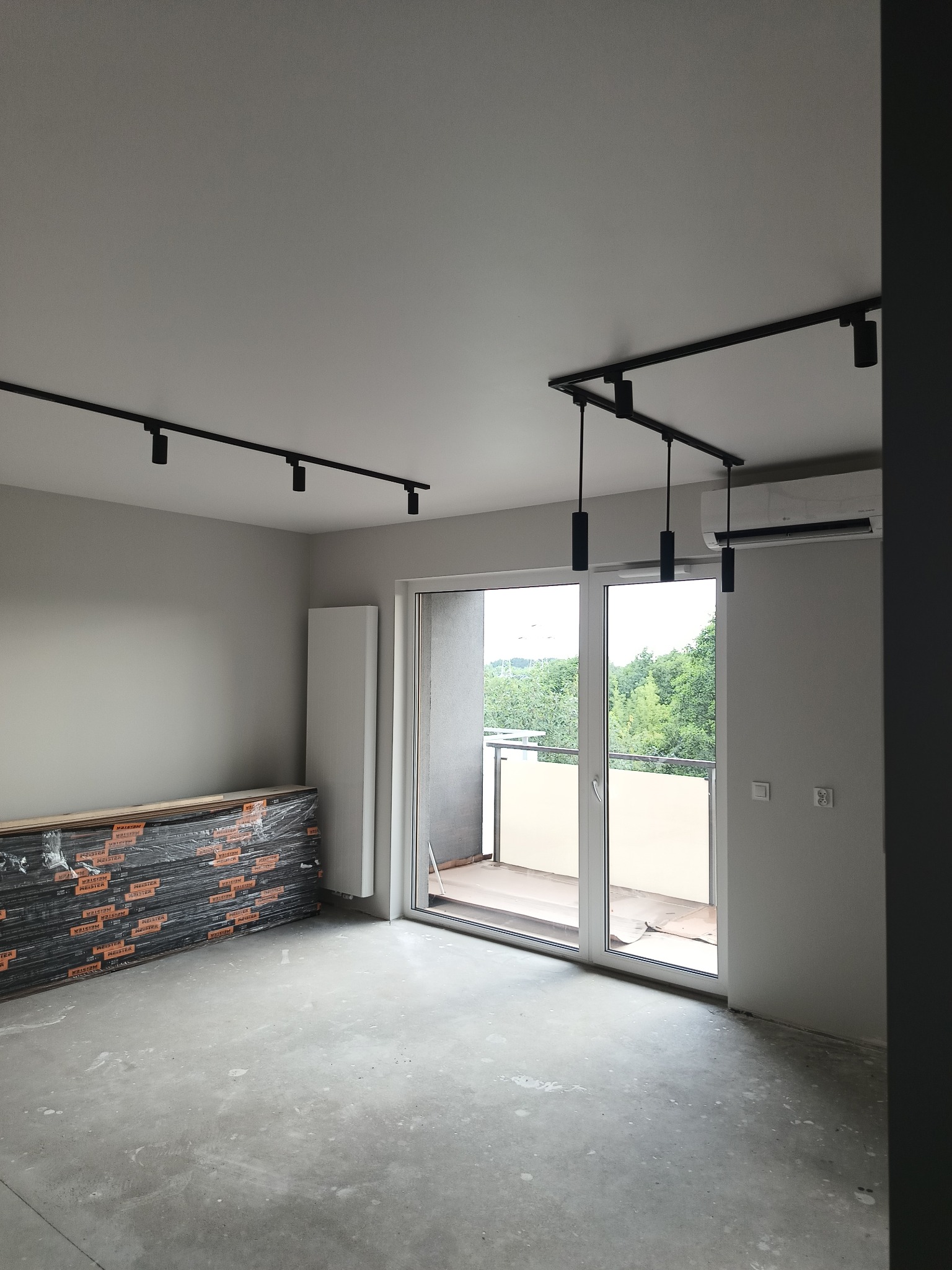 Surowe wnętrze mieszkania w trakcie remontu: widoczne panele podłogowe w folii, surowy beton na podłodze, czarne szyny oświetleniowe na suficie i klimatyzator ścienny, z wyjściem na balkon...