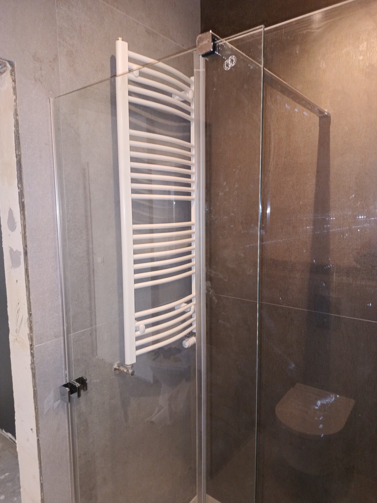 Szklana kabina prysznicowa z grzejnikiem drabinkowym w trakcie montażu, widoczne niedokończone wykończenia ściany