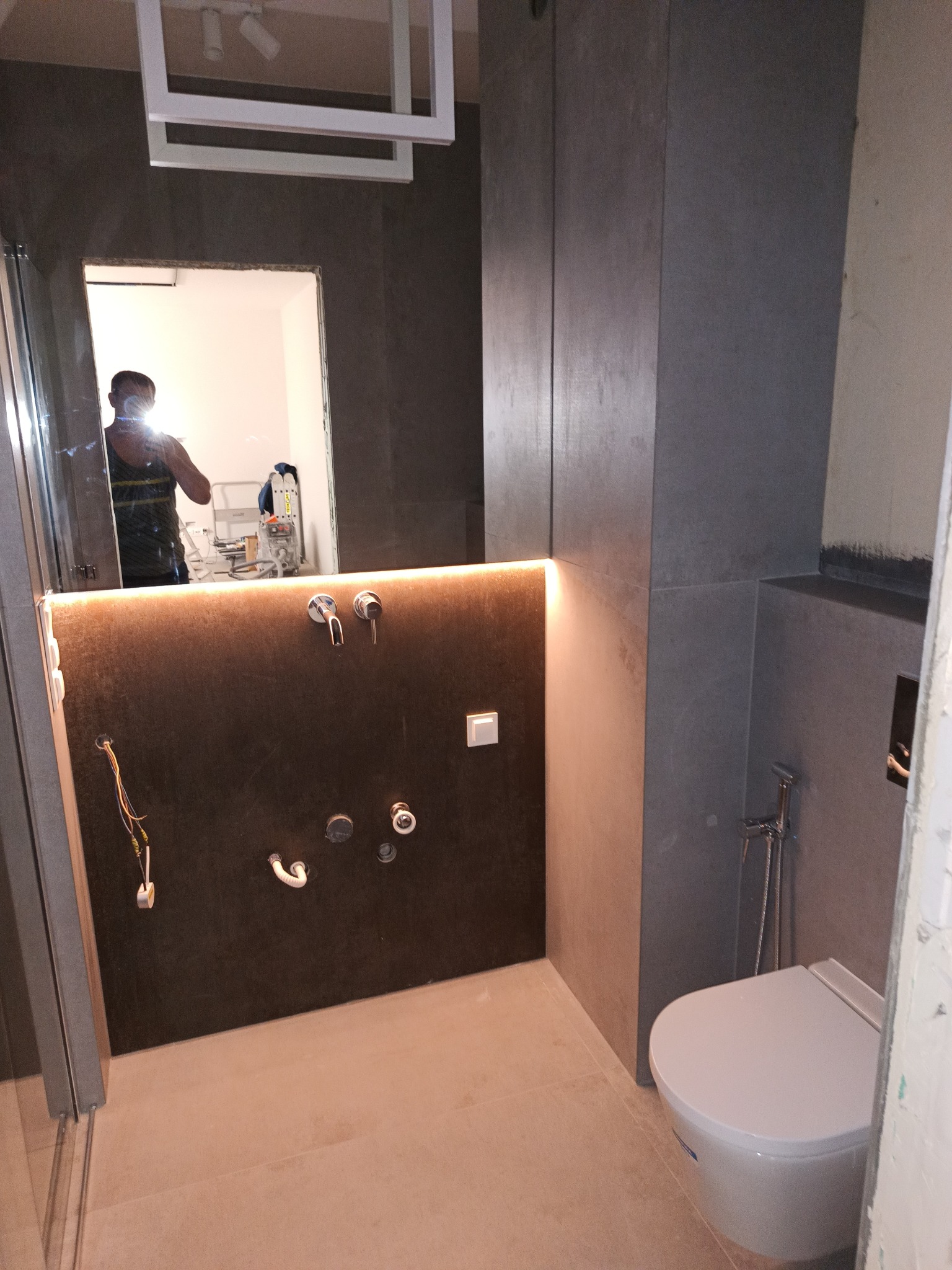 Wnętrze łazienki w trakcie prac wykończeniowych, widoczne szare płytki na ścianach, podświetlona wnęka z wyprowadzonymi instalacjami wodnymi, toaleta wisząca oraz odbicie osoby w lustrze.
