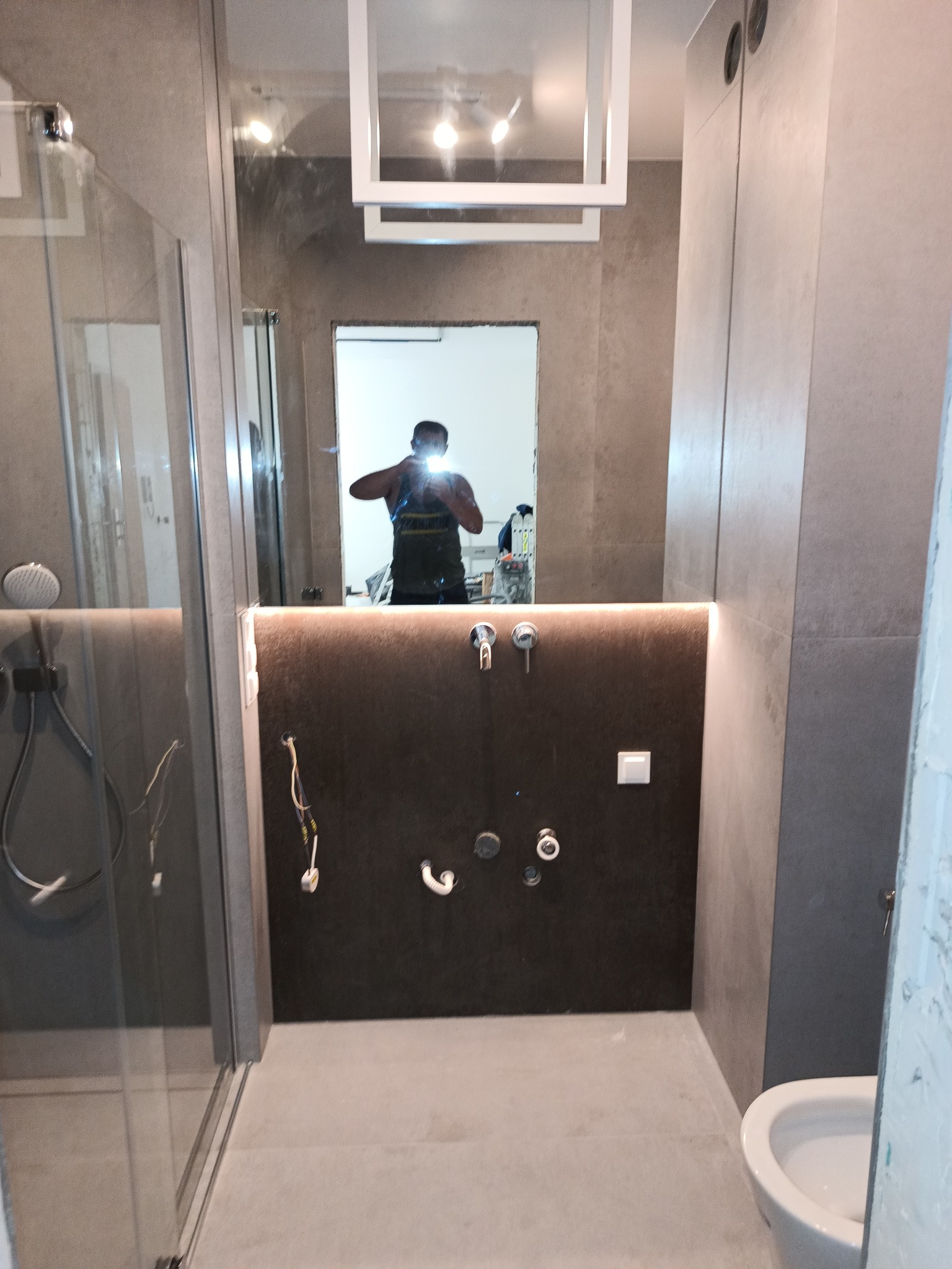 Szara łazienka w trakcie prac wykończeniowych, widoczne przygotowane przyłącza wodno-kanalizacyjne, częściowo zamontowana kabina prysznicowa i toaleta, oraz odbicie osoby robiącej zdjęcie w lustrze.