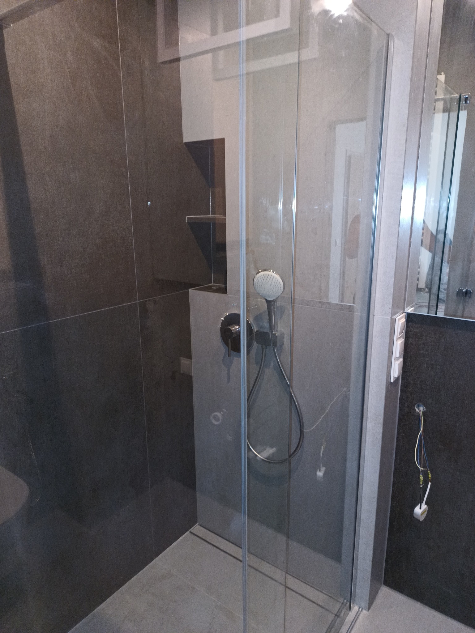 Nowoczesna kabina prysznicowa z ciemnymi, matowymi płytkami, deszczownicą i szklanymi drzwiami, widoczne niedokończone podłączenia elektryczne.