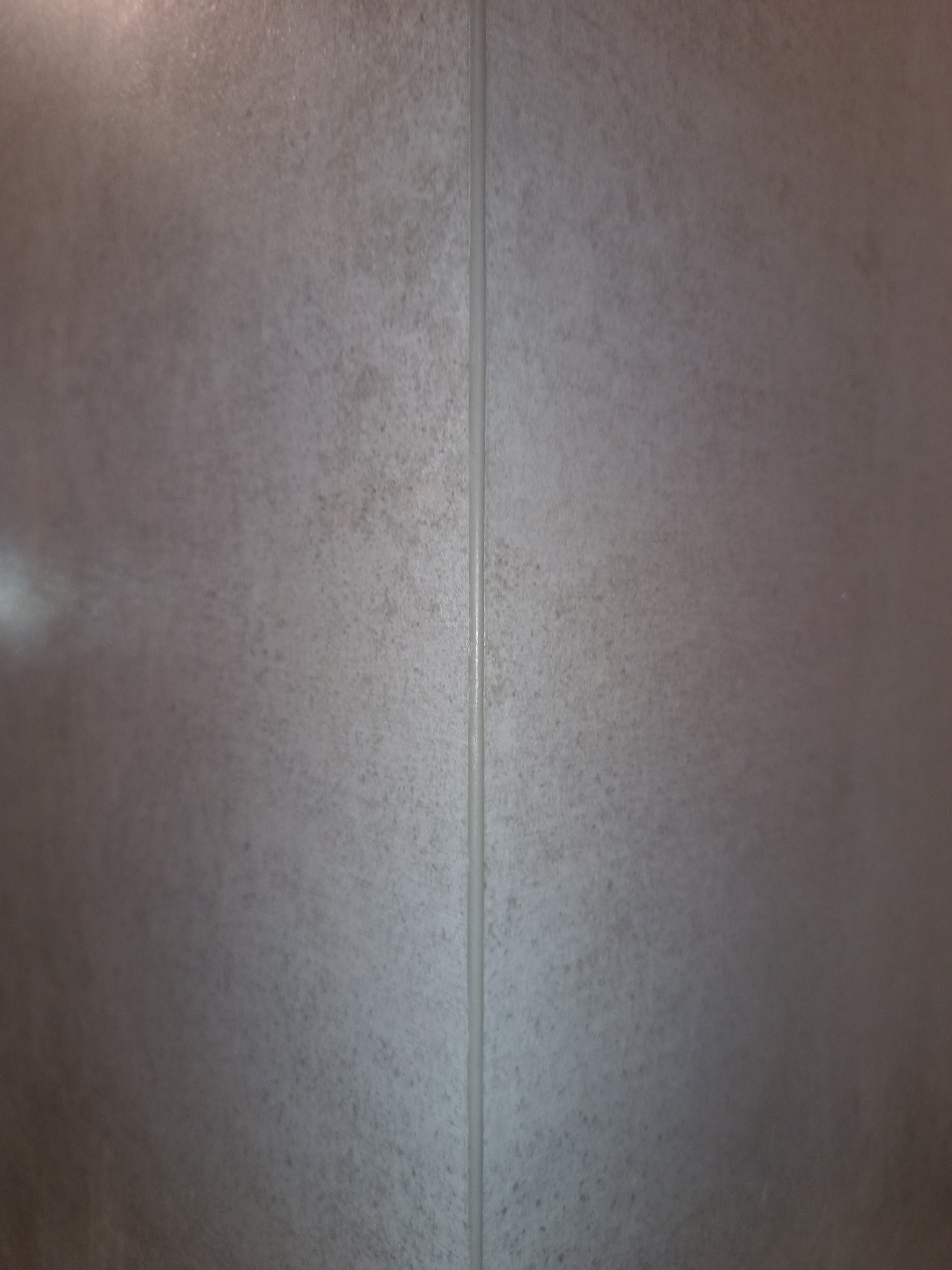 Fragment ściany wyłożonej szarą, matową płytką ceramiczną z widoczną fugą pośrodku, oświetlenie z lewej strony.