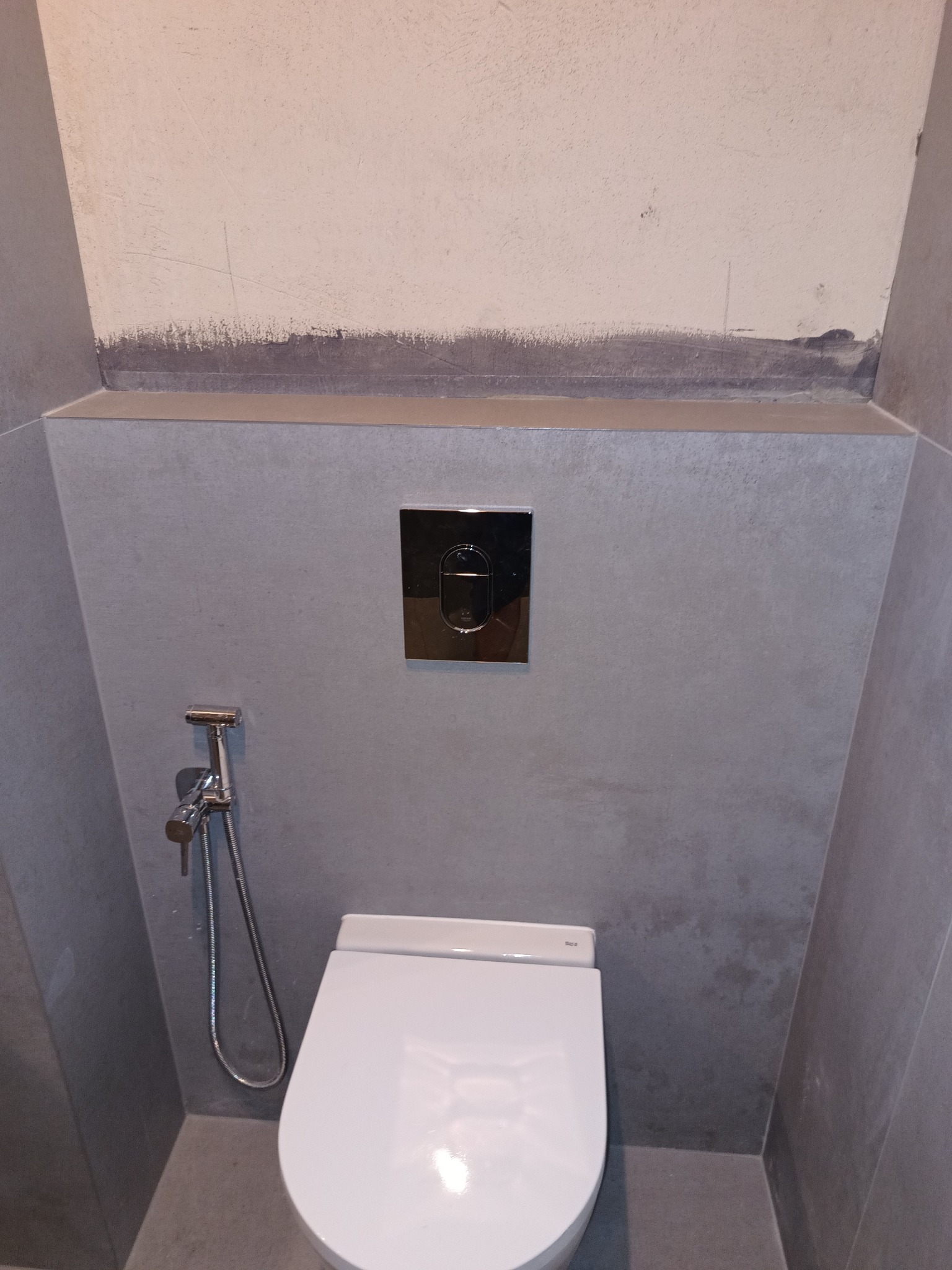 Zawieszona miska WC z chromowanym przyciskiem spłukującym i bidetką natryskową na tle szarej ściany z widocznymi śladami po remoncie.