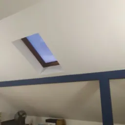 Okno dachowe z drewnianą ramą w białej ścianie poddasza, widok na błękitne niebo, poniżej ściana pomalowana na biało z niebieską ramą dekoracyjną.