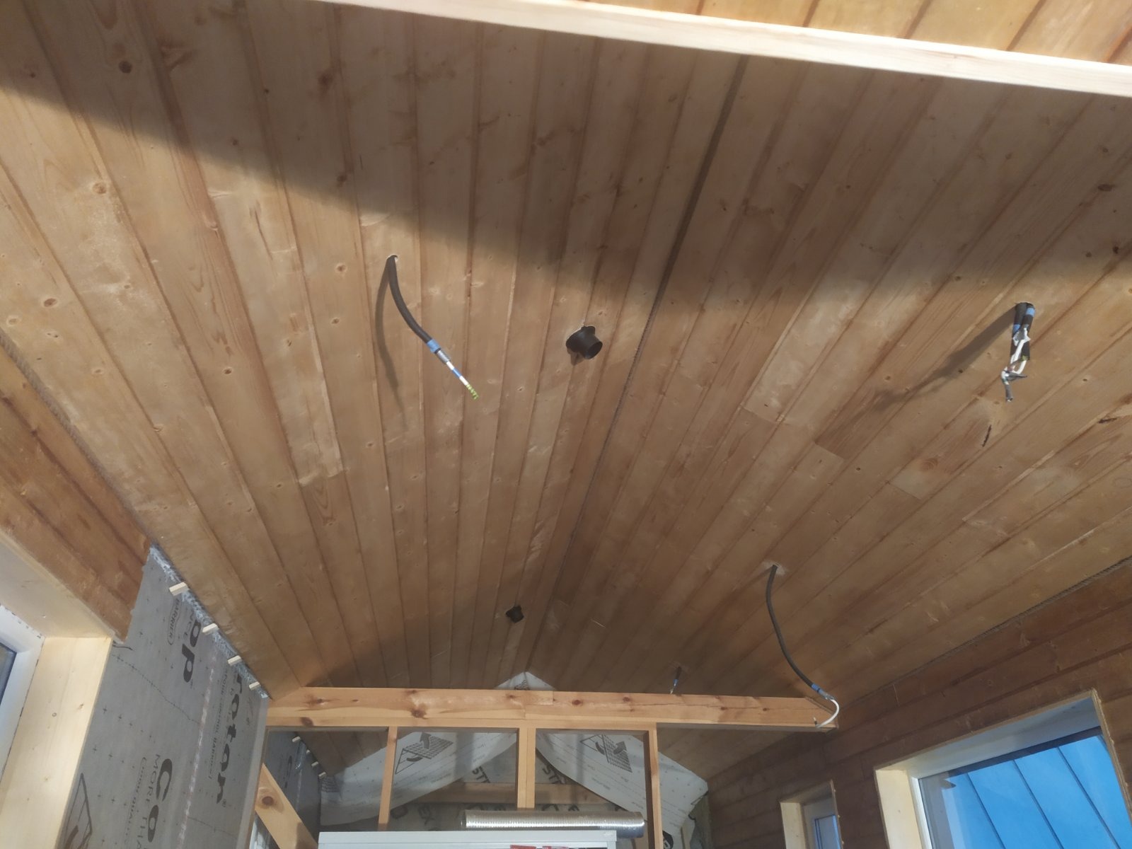 Drewniany sufit z widocznymi kablami elektrycznymi i otworami, częściowo wykończony, z widoczną konstrukcją i oknami w tle.