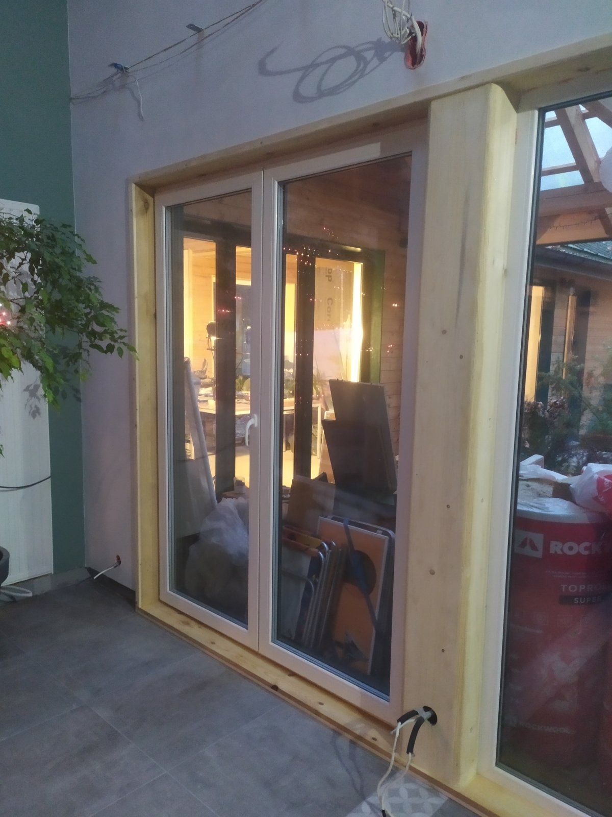 Montaż okna tarasowego z drewnianą ramą, widoczne materiały budowlane i narzędzia wewnątrz pomieszczenia.