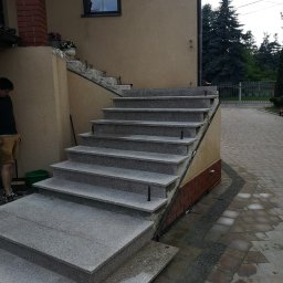 Remonty A&P - Zewnętrzne schody z szarego granitu prowadzące do domu, widoczne elementy wykończenia cegłą klinkierową, obok stoi mężczyzna z miotłą i zielonym wiadrem.