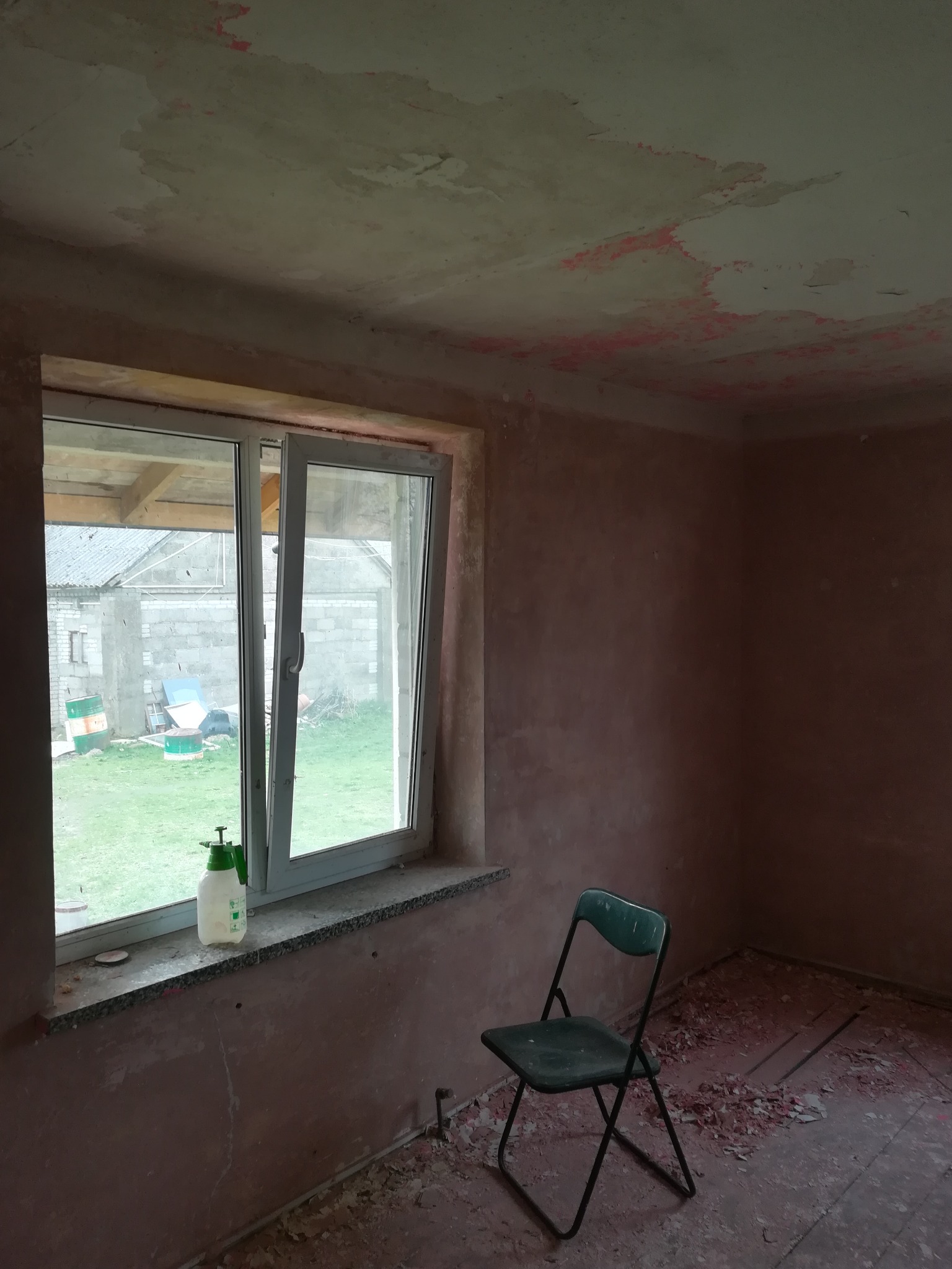 Pokój w trakcie przygotowania do remontu: surowe ściany z resztkami starej farby, okno z widokiem na podwórze, plastikowy opryskiwacz na parapecie, zielone krzesło i resztki gruzu na podłodze.