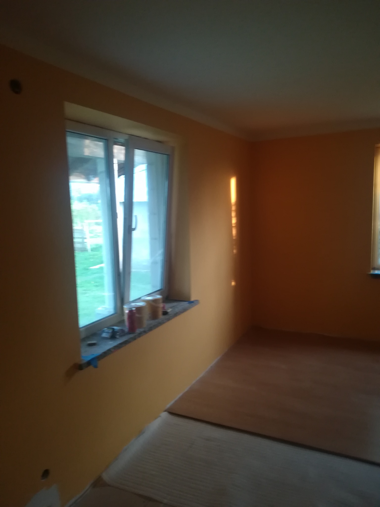 Pokój po malowaniu ścian na żółto-pomarańczowy kolor, widoczne puszki z farbą na parapecie okna oraz częściowo położona podłoga.