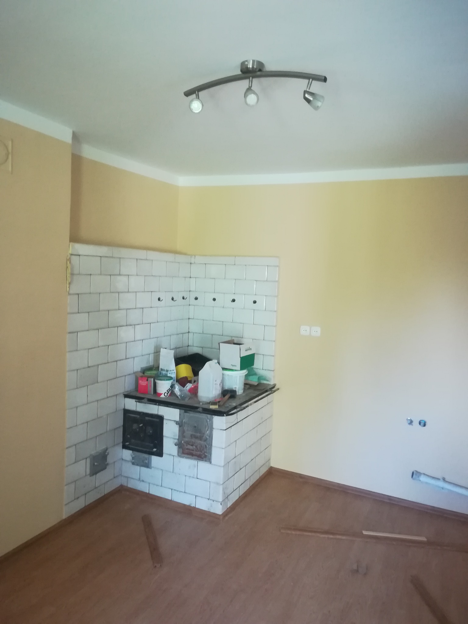 Fragment kuchni w trakcie prac remontowych: piec kaflowy obłożony białymi płytkami, widoczne materiały budowlane i narzędzia, surowe ściany w odcieniach beżu, drewnopodobna podłoga z listwami.