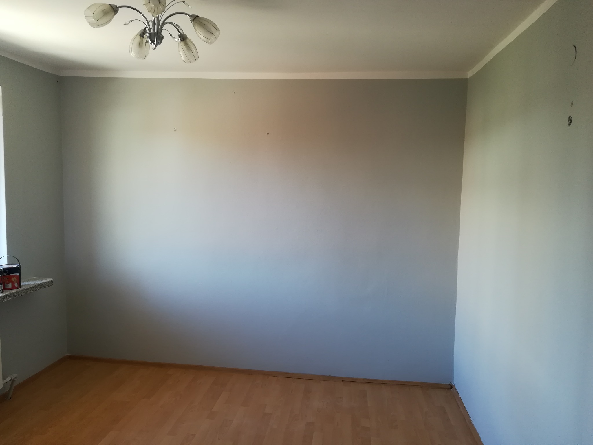 Pomalowane na szaro ściany w pustym pokoju z parkietem, widoczne otwory po kołkach i lampa sufitowa.