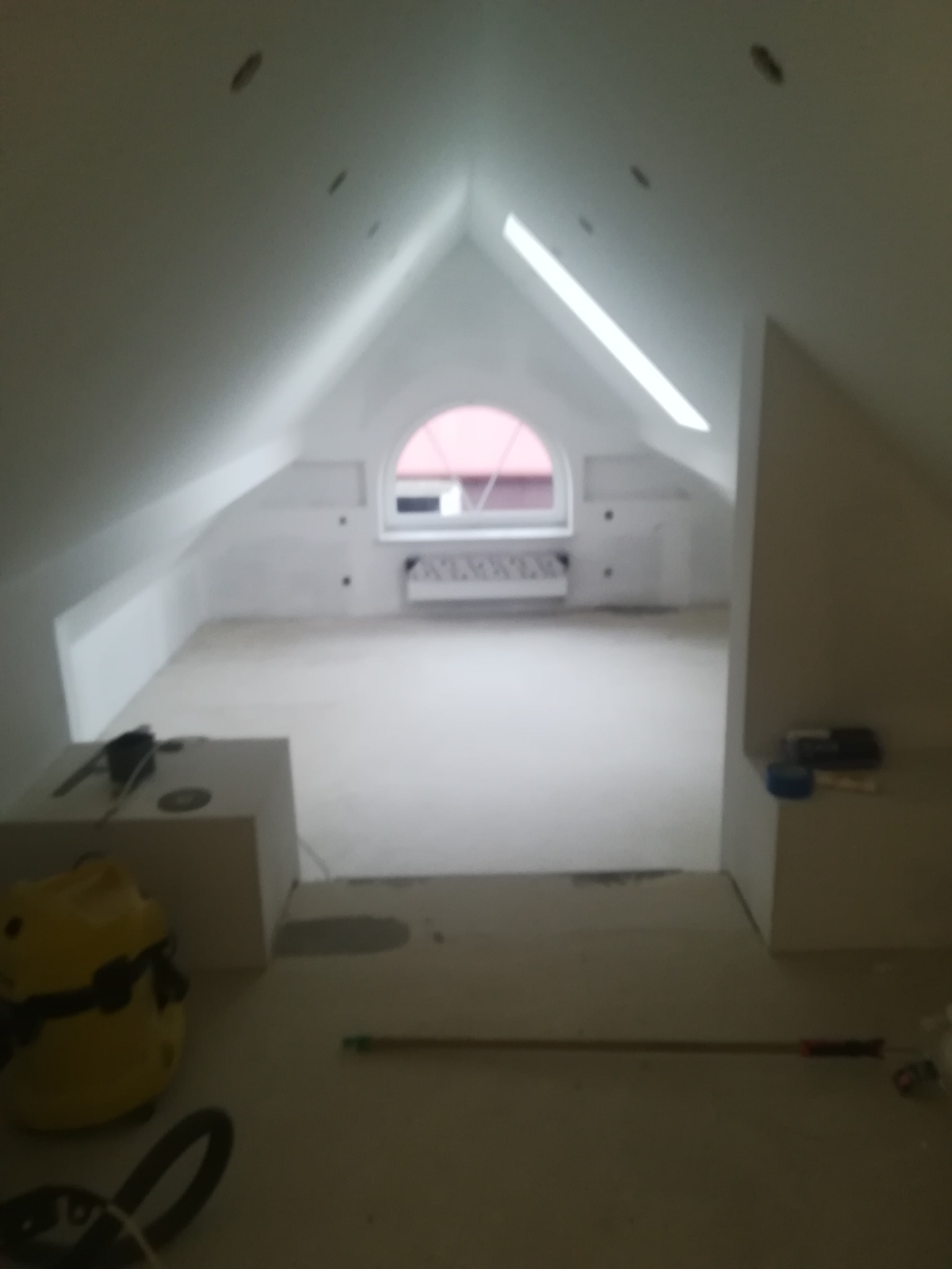 Poddasze w trakcie prac przygotowawczych do wykończenia, widoczne okno dachowe, grzejnik, narzędzia i materiały budowlane, ściany przygotowane do malowania.