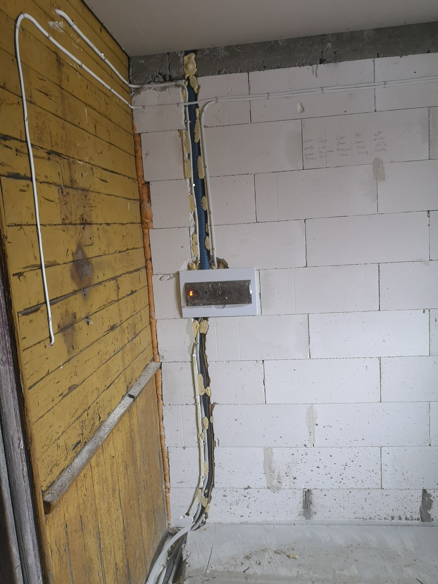 Instalacja elektryczna w trakcie montażu: przewody w peszlach mocowane pianką montażową do ściany z bloczków, obok fragmentu drewnianej ściany z żółtą farbą. Na ścianie widoczne odręczne notatki.