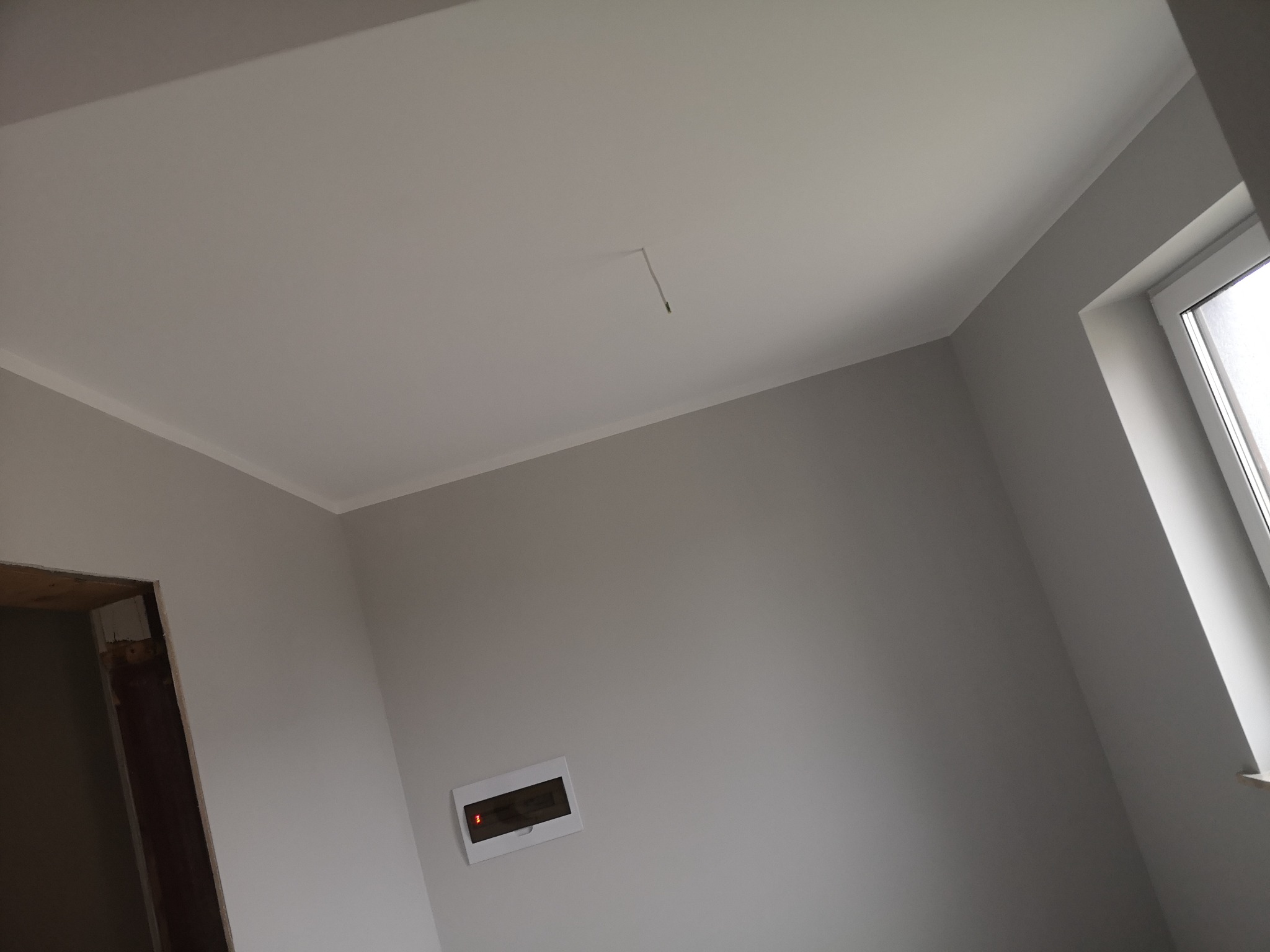 Wnętrze pokoju po remoncie: świeżo pomalowane ściany na szaro, biały sufit z listwą, okno z białymi ramami, widoczna skrzynka elektryczna i ościeżnica drzwi.