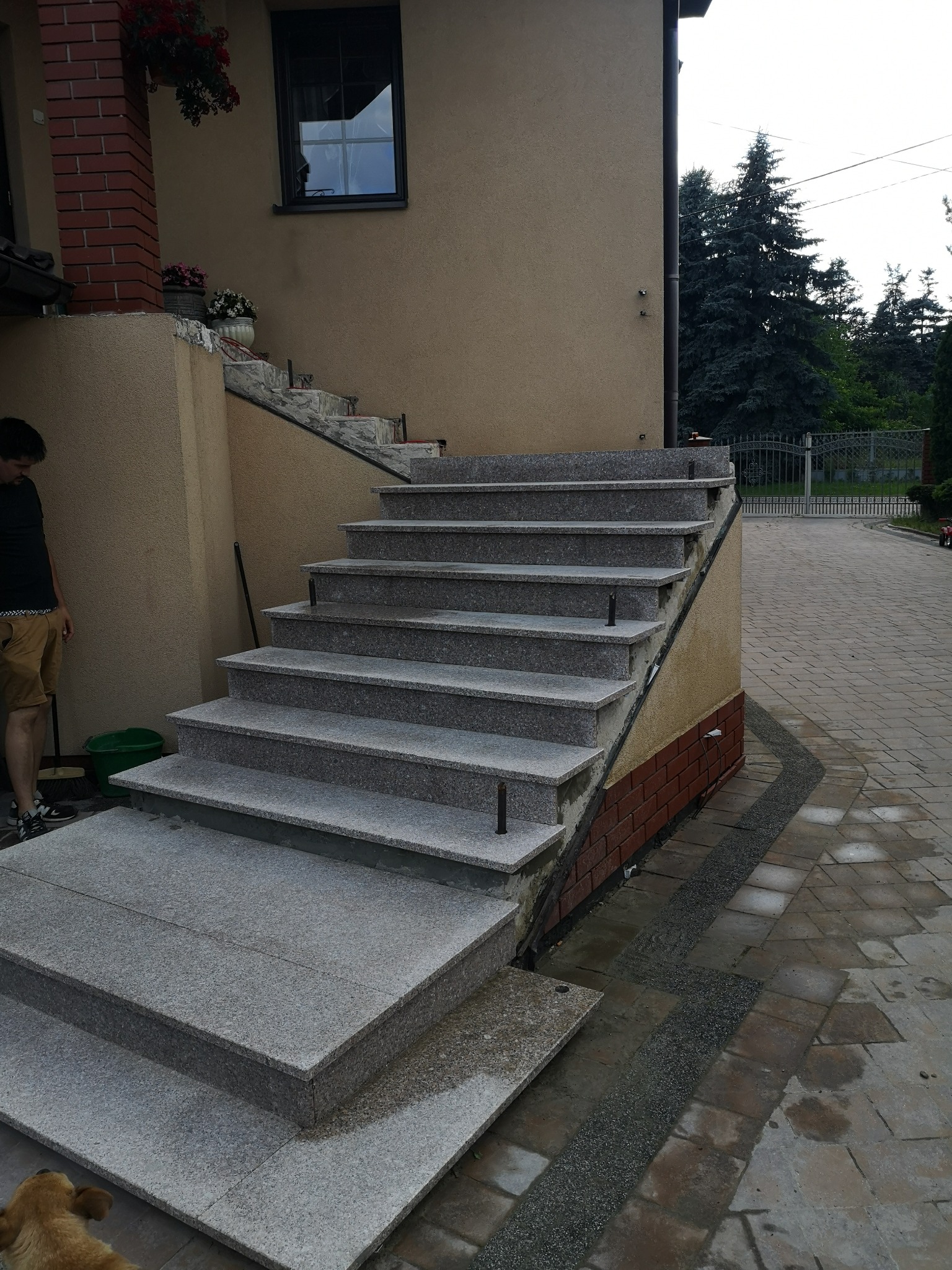 Zewnętrzne schody z szarego granitu prowadzące do domu, widoczne elementy wykończenia cegłą klinkierową, obok stoi mężczyzna z miotłą i zielonym wiadrem.