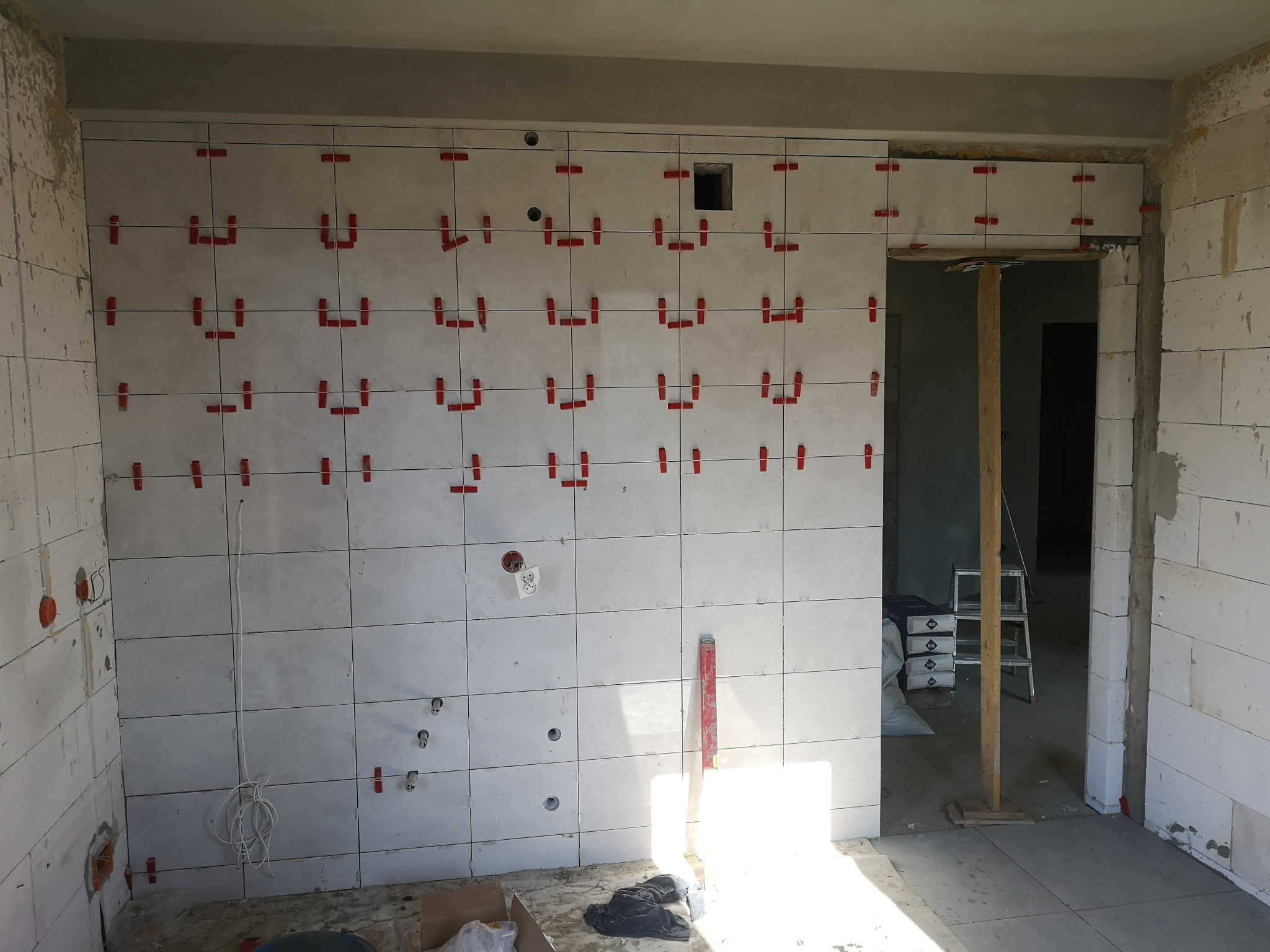 Ściana w trakcie układania białych płytek, z widocznymi czerwonymi krzyżykami dystansowymi, otwory na instalacje, surowy stan wykończenia, widok na sąsiednie pomieszczenie z drabiną i workami zaprawy.