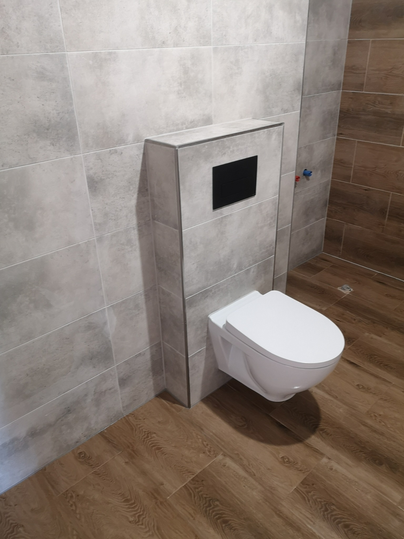 Nowoczesna toaleta podwieszana z szarą zabudową stelaża i czarnym panelem sterującym, wykończona płytkami imitującymi beton i drewno.